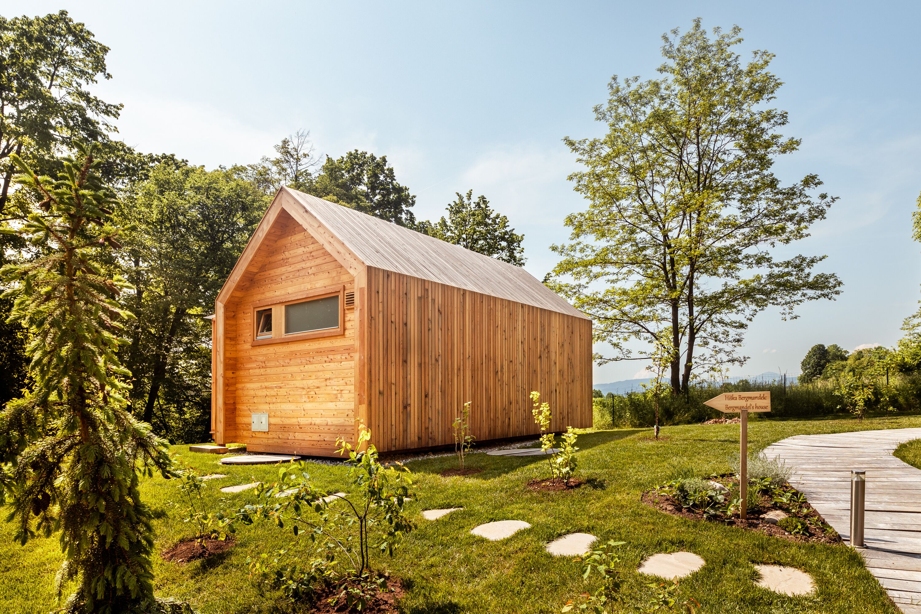 A wooden cabin with a pitched roof, surrounded by a well-maintained garden with stepping stones and a sign indicating a nearby location.