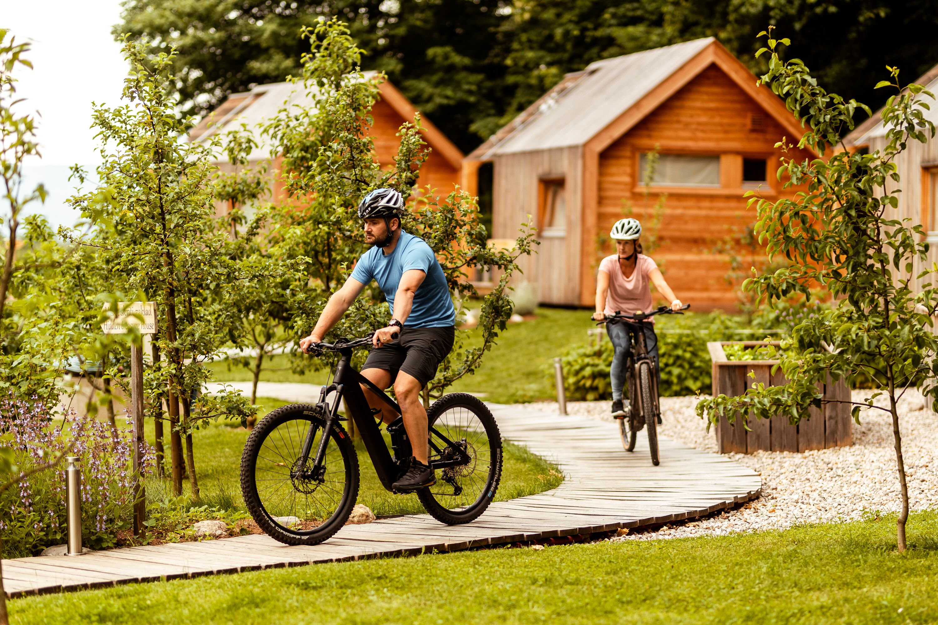 Dois ciclistas em uma trilha de madeira com cabanas de madeira ao fundo.