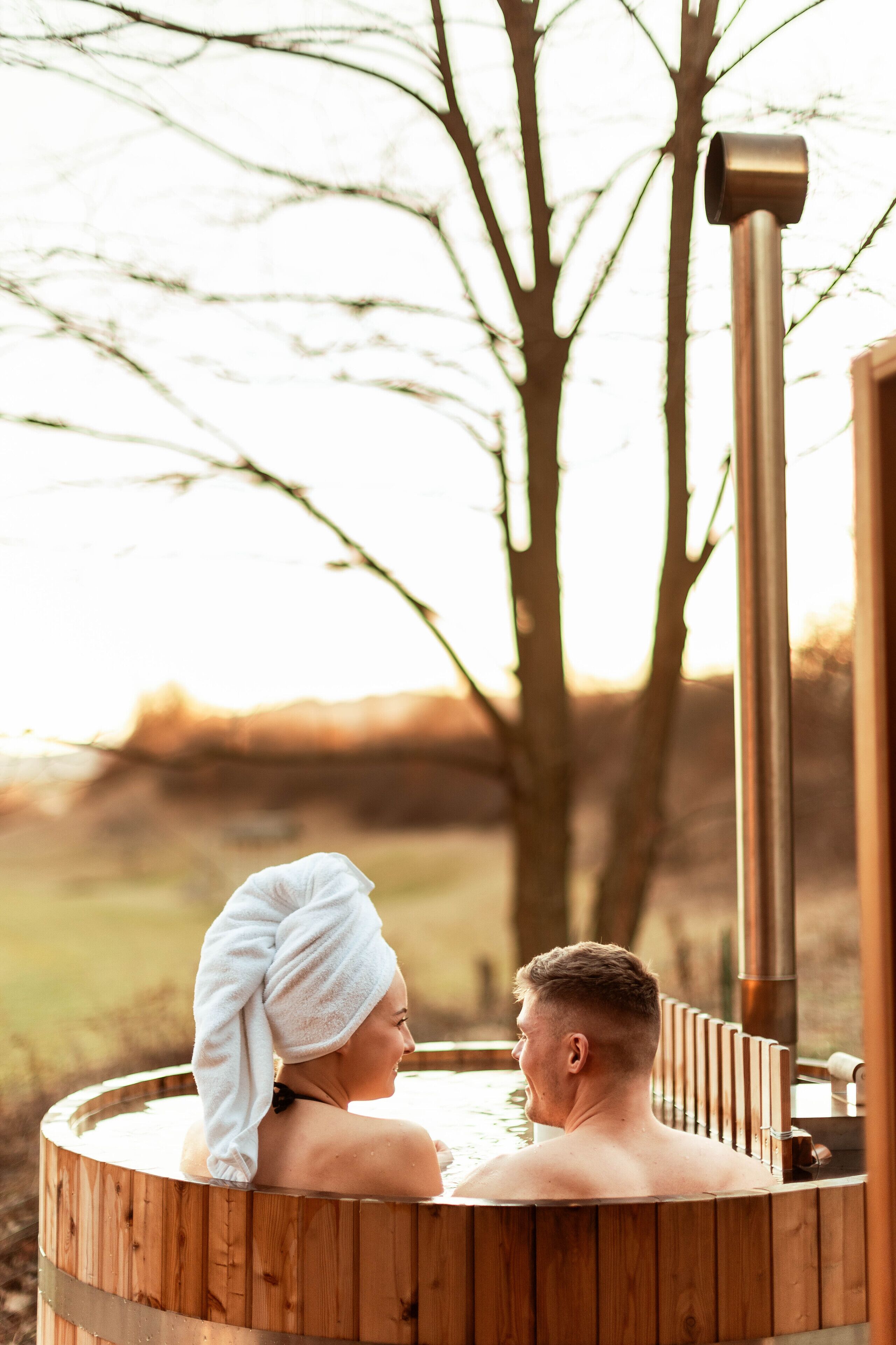 A wooden hot tub with a person and a towel, surrounded by trees and a wooden structure.