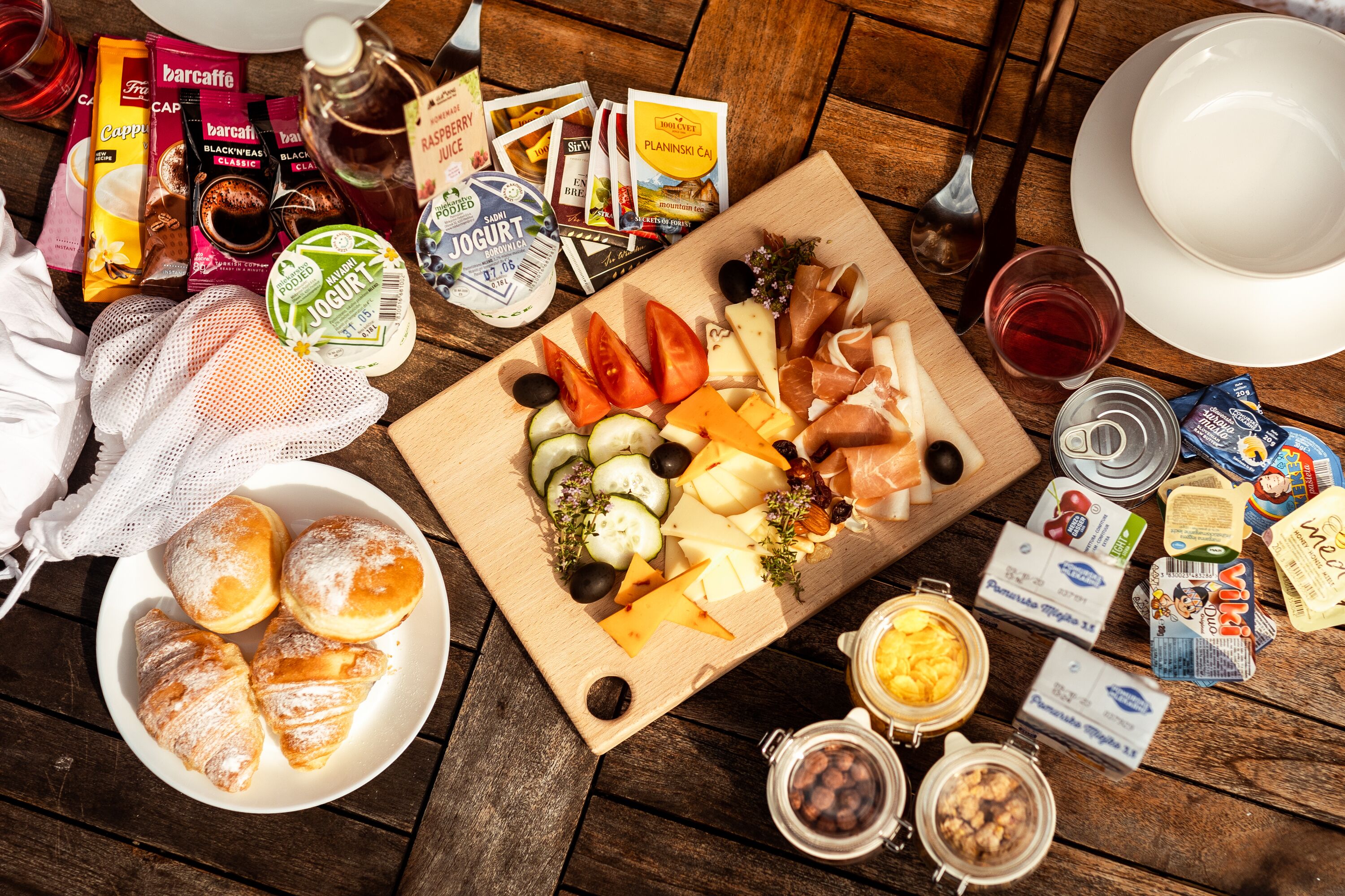 A wooden table with a variety of food items, including cheese, olives, and pastries, along with several packaged snacks and condiments.