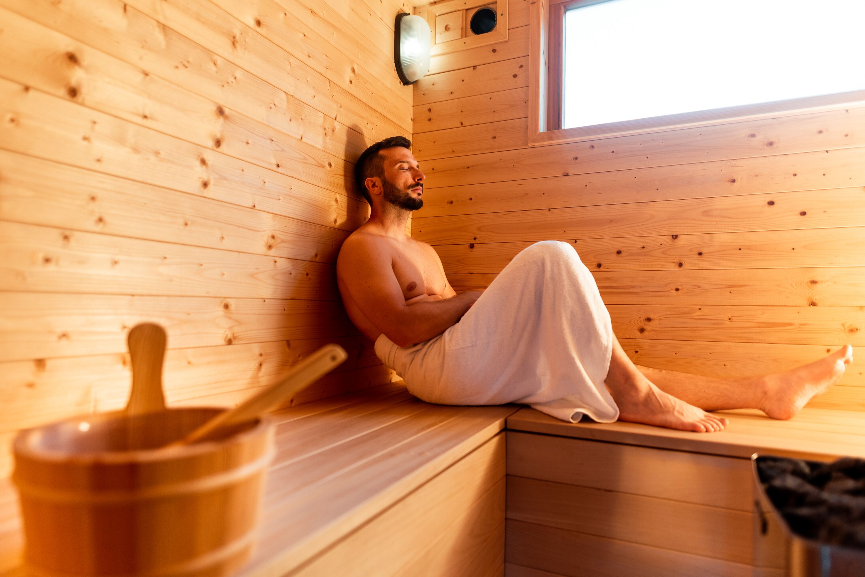 A person sitting in a wooden sauna with a wooden bench and a small wooden bowl.