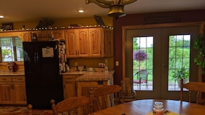 A kitchen with wooden cabinets, a black refrigerator, and a dining table with chairs. There is a window with a view of greenery outside.