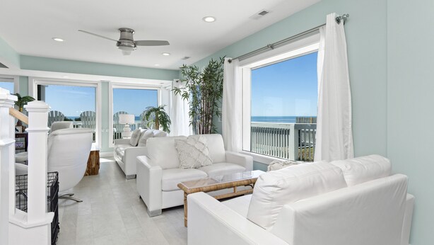 A spacious living area with white furniture, a large window with a view of the ocean, and a ceiling fan.