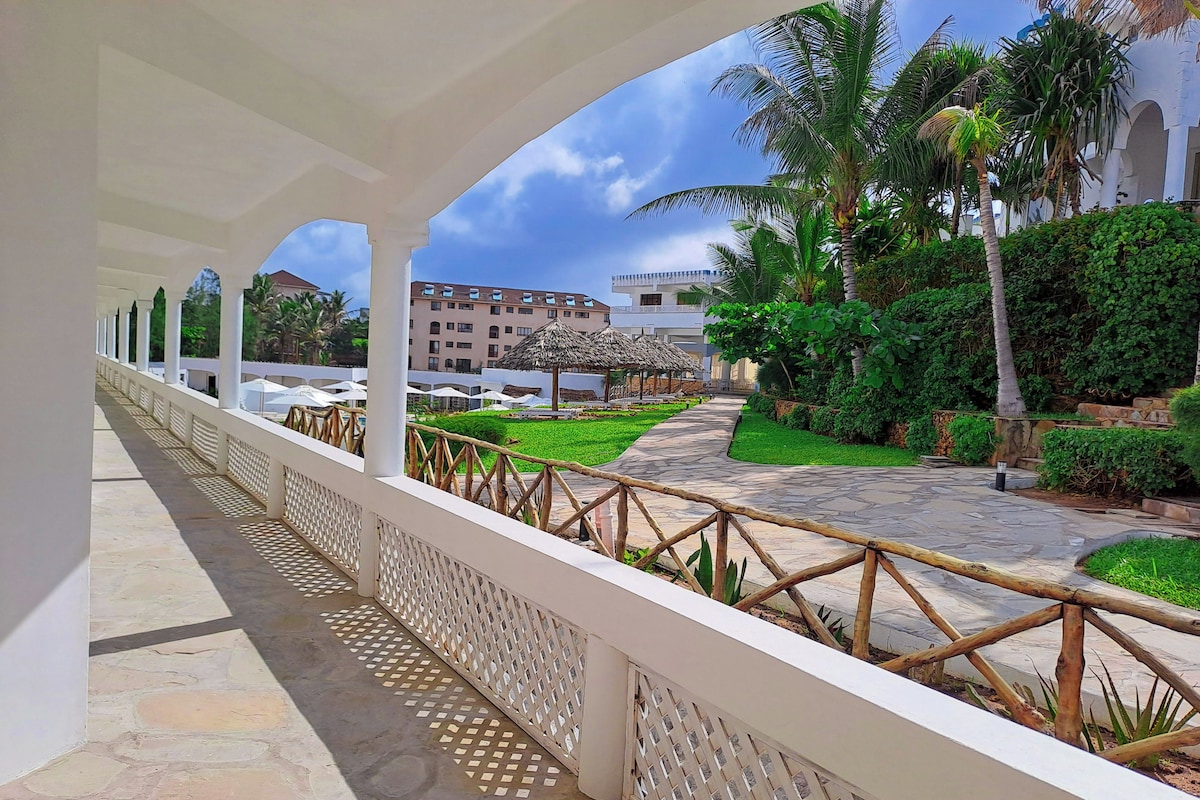 A covered walkway with a white railing, leading to a courtyard with a thatched-roof gazebo, palm trees, and a building in the background.