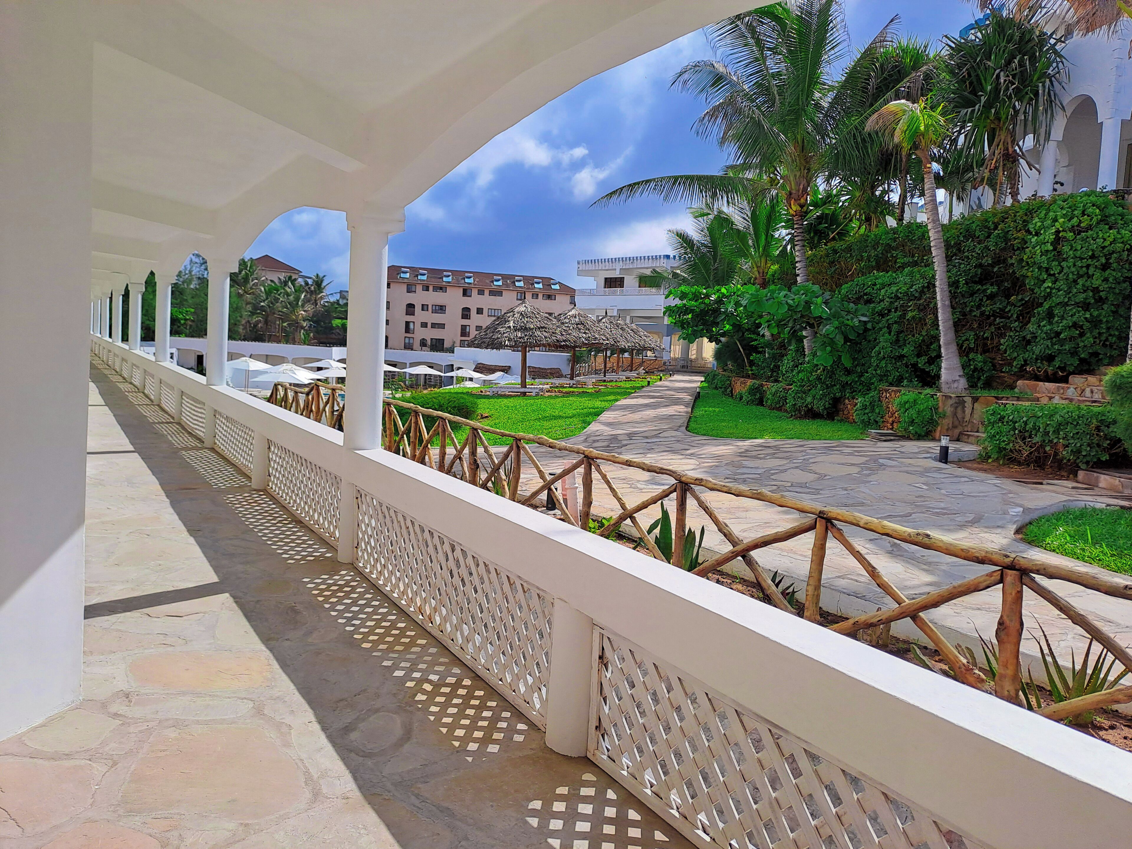 A covered walkway with a white railing, leading to a courtyard with a thatched-roof gazebo, palm trees, and a building in the background.