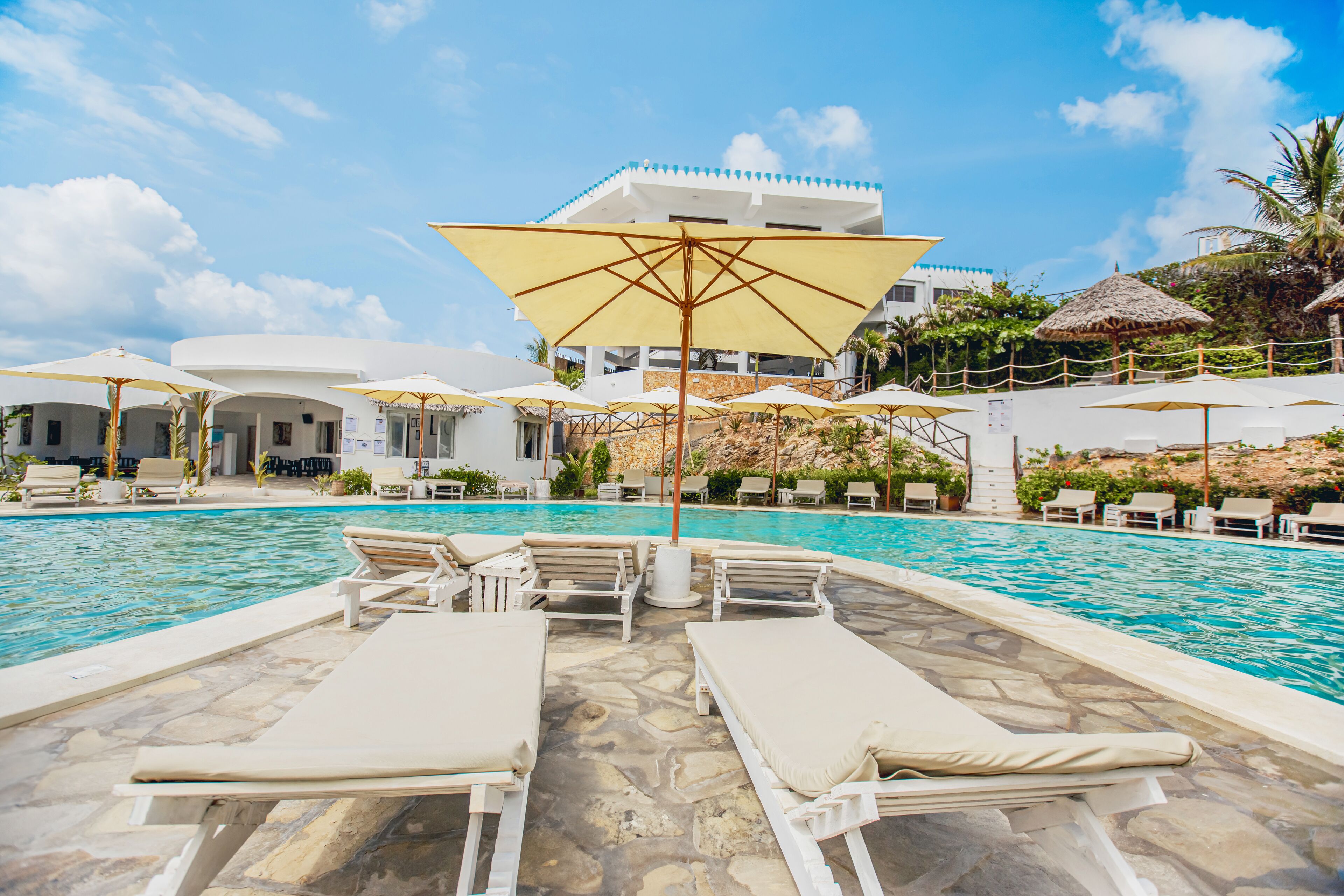 A pool area with lounge chairs and umbrellas, surrounded by a stone deck and a building with a white facade and blue roof.