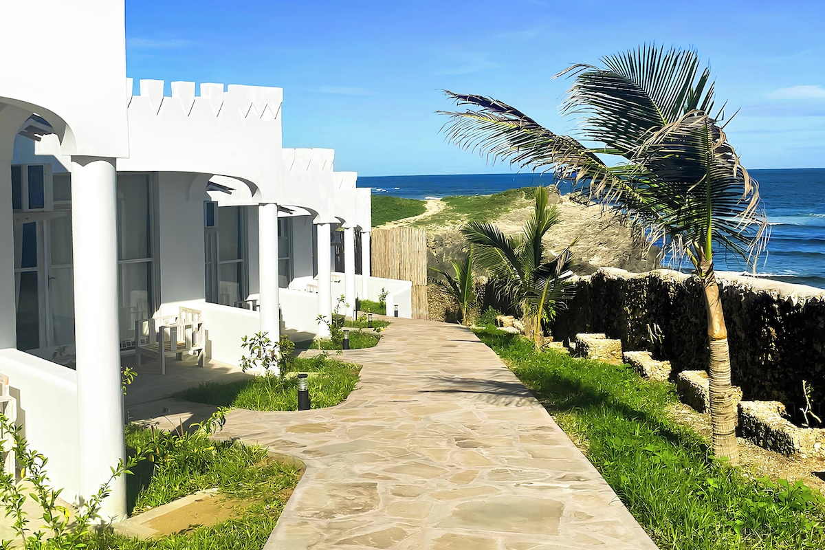A pathway leading to a white building with columns, a palm tree, and a view of the ocean.