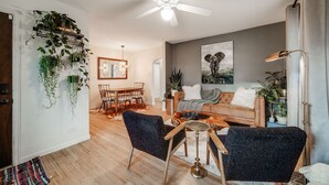 A modern living room with a ceiling fan, a dining area with a wooden table and chairs, and a sofa with a throw blanket. - Charming 3 Bedroom 1940s Farm House (Lakewood)
