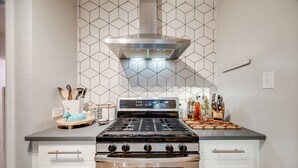 A kitchen with a gas stove, oven, and stainless steel range hood. There are cabinets with handles, a countertop with various kitchen items, and a backsplash with a geometric pattern. - Charming 3 Bedroom 1940s Farm House (Lakewood)