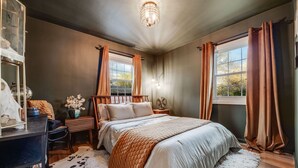 A bedroom with a bed, a desk, a chair, a nightstand, a lamp, and a window with curtains. - Charming 3 Bedroom 1940s Farm House (Lakewood)