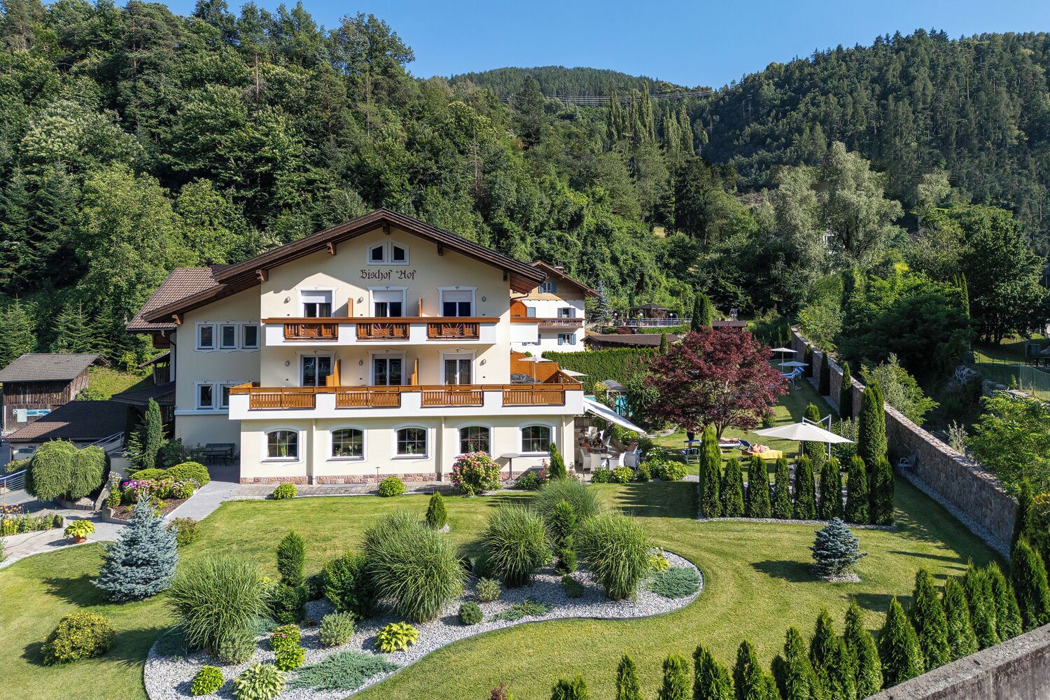 A multi-story hotel with balconies, surrounded by a well-maintained garden and forested hills.