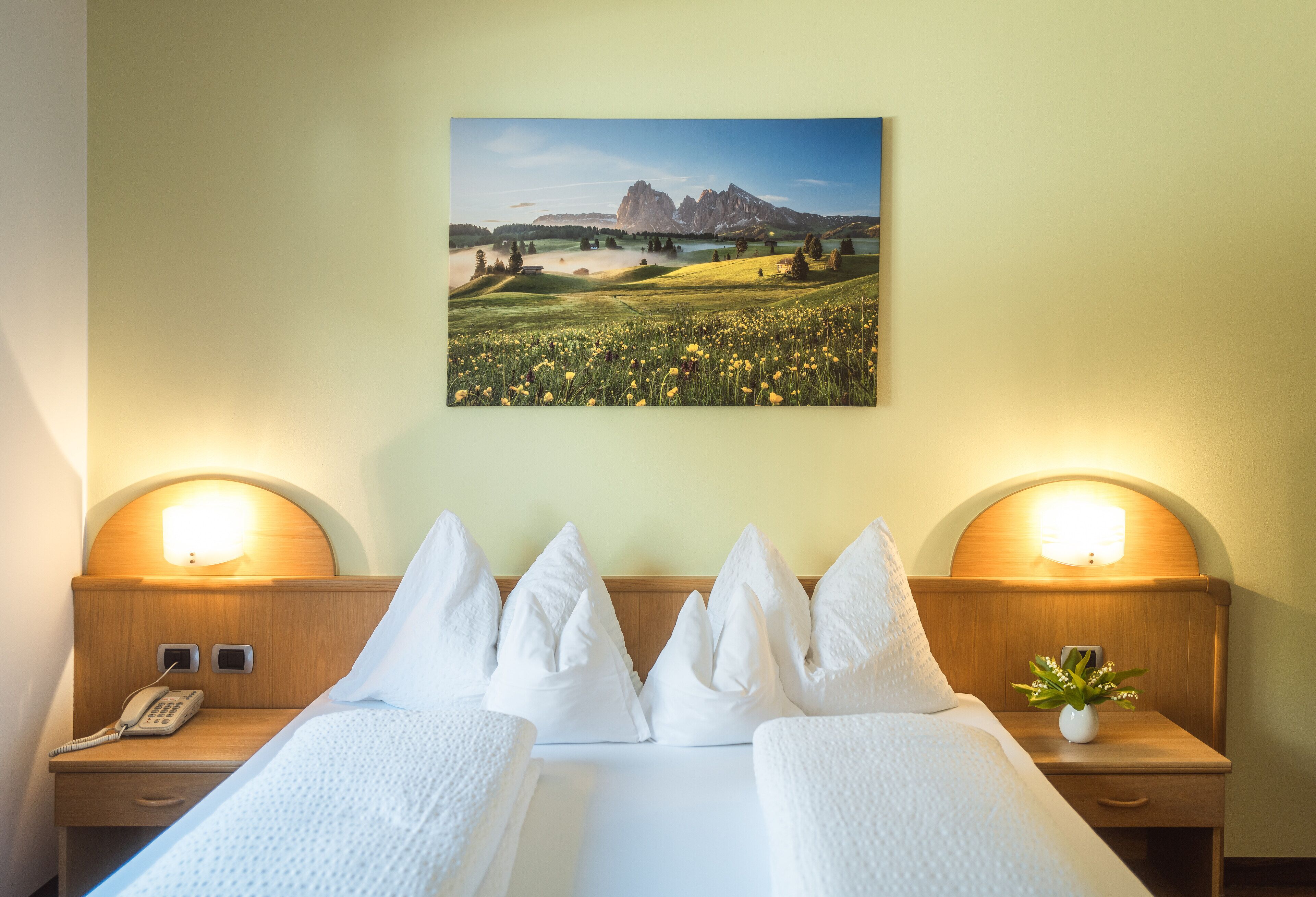 A hotel room with a bed, two bedside tables, a nightstand, a telephone, and a wall-mounted painting of a landscape.