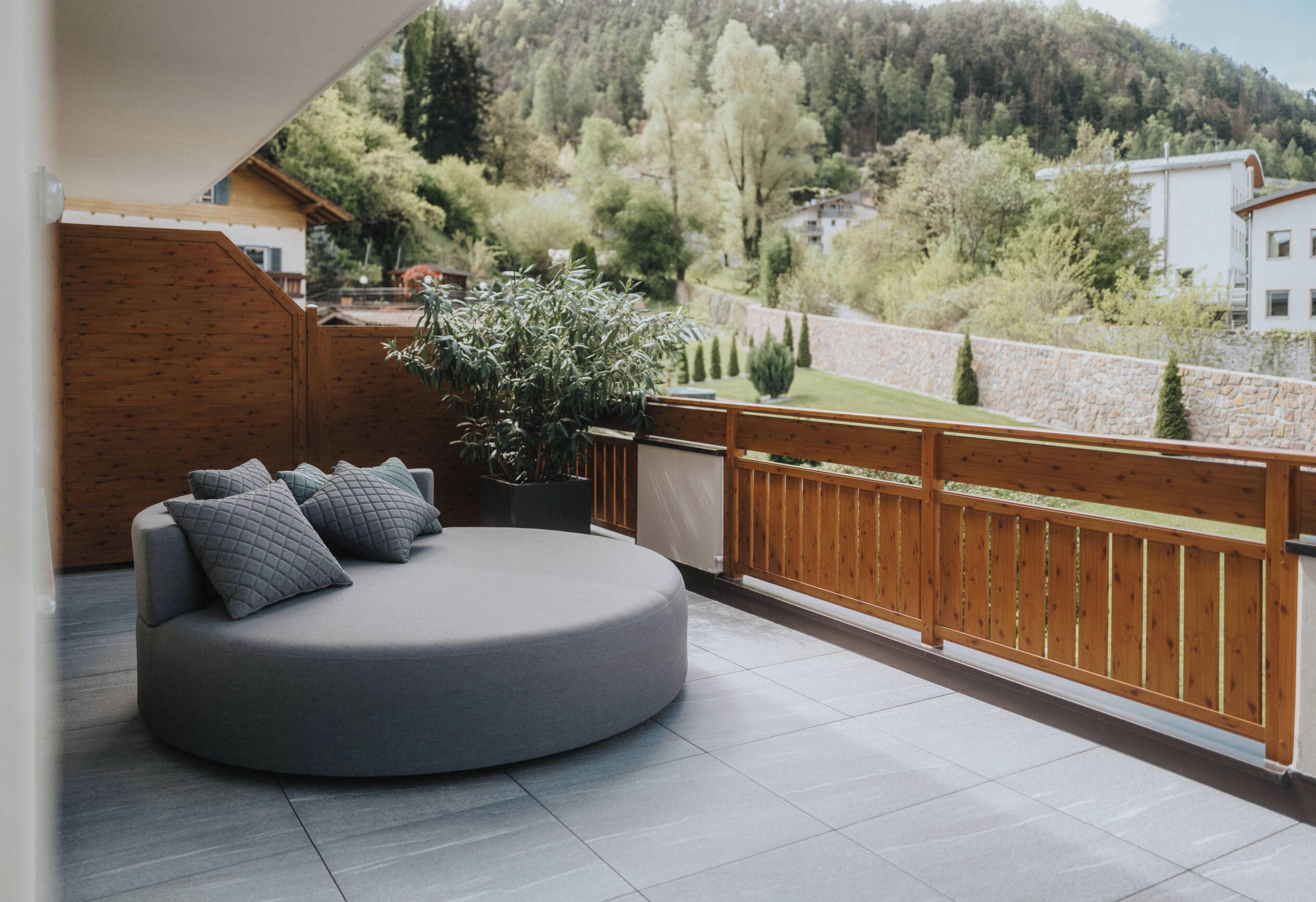 A modern outdoor seating area with a round grey sofa and cushions, a wooden fence, and a view of a hilly landscape.