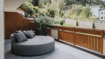 A modern outdoor seating area with a round grey sofa and cushions, a wooden fence, and a view of a hilly landscape.