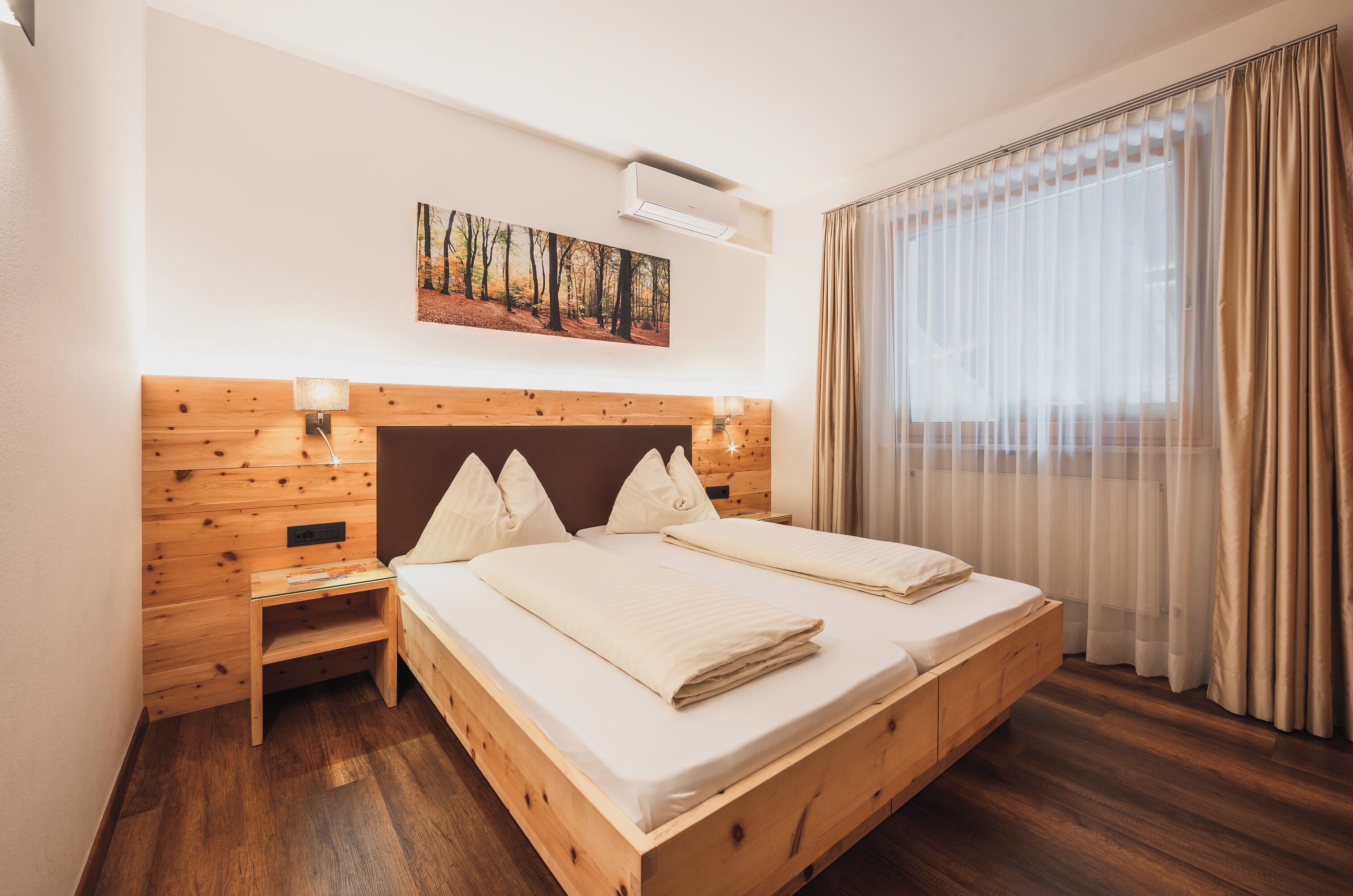 Una cama doble con cabecera de madera, una mesita de noche, aire acondicionado instalado en la pared y un ventanal con cortinas traslúcidas.