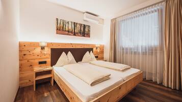 Una cama doble con cabecera de madera, una mesita de noche, aire acondicionado instalado en la pared y un ventanal con cortinas traslúcidas.