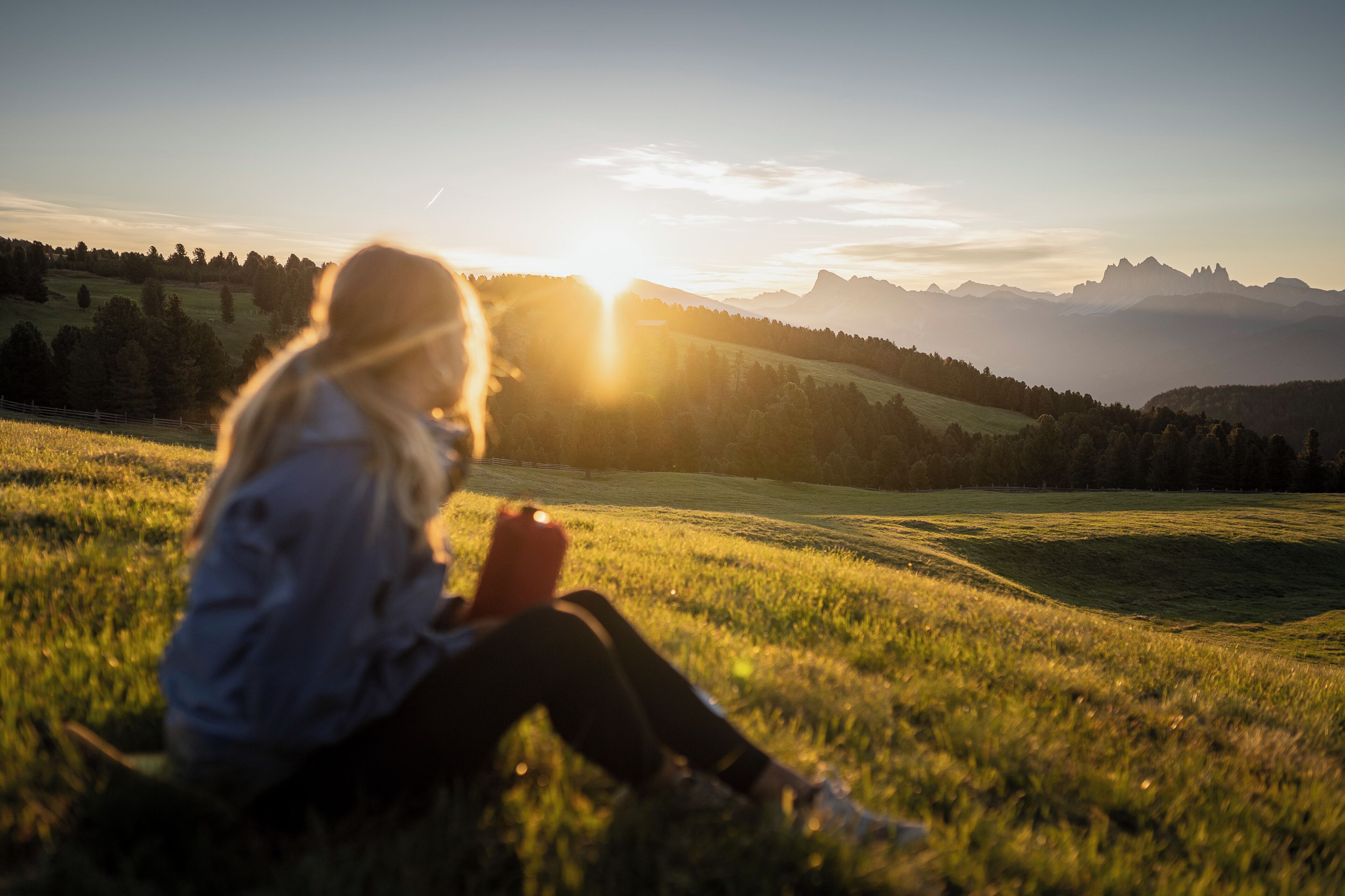 En person sitter på gräset med en vacker utsikt över berg och en solnedgång i bakgrunden.