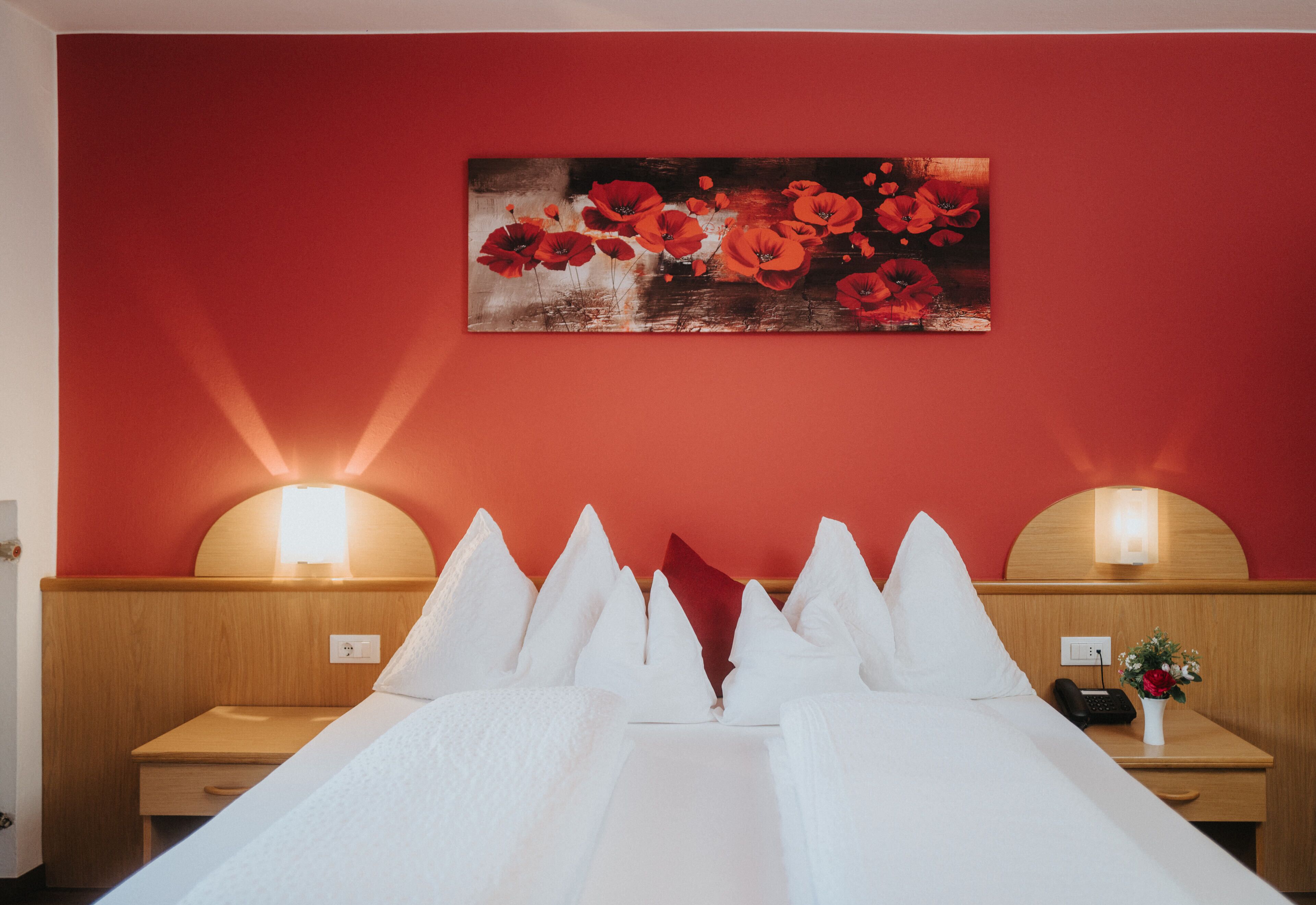 Une chambre d’hôtel avec un mur rouge, un lit recouvert de draps blancs, deux tables de chevet avec des lampes et un tableau représentant des fleurs rouges.