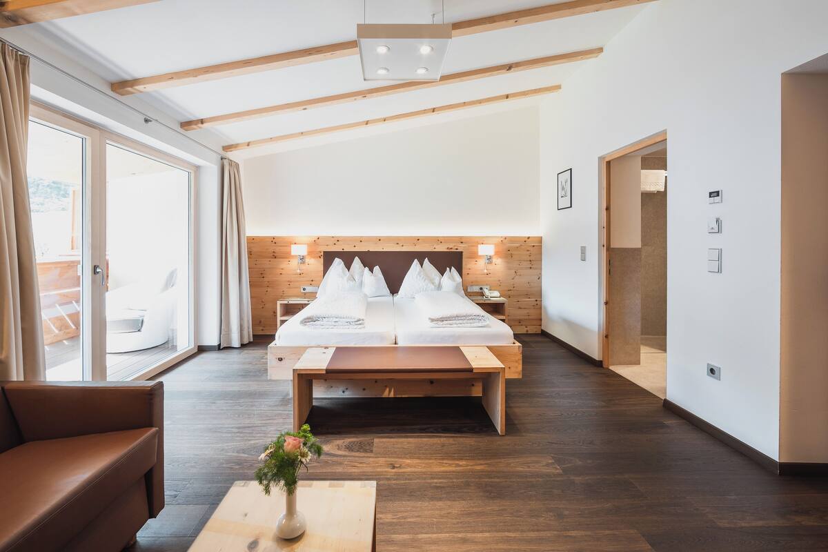 Een moderne hotelkamer met een houten plafond, een groot bed met witte lakens, een houten bankje en uitzicht door grote ramen.