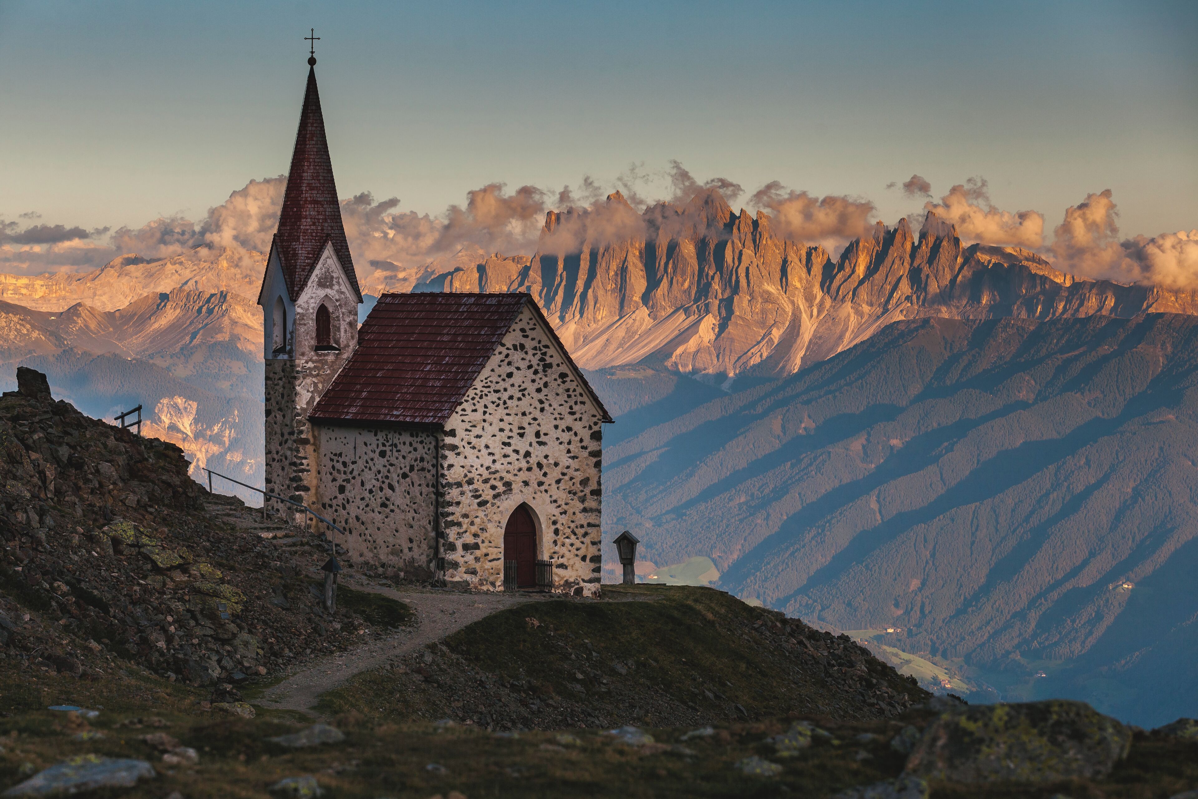 A small stone church with a steeply pitched roof and a cross on top, surrounded by rugged mountains.