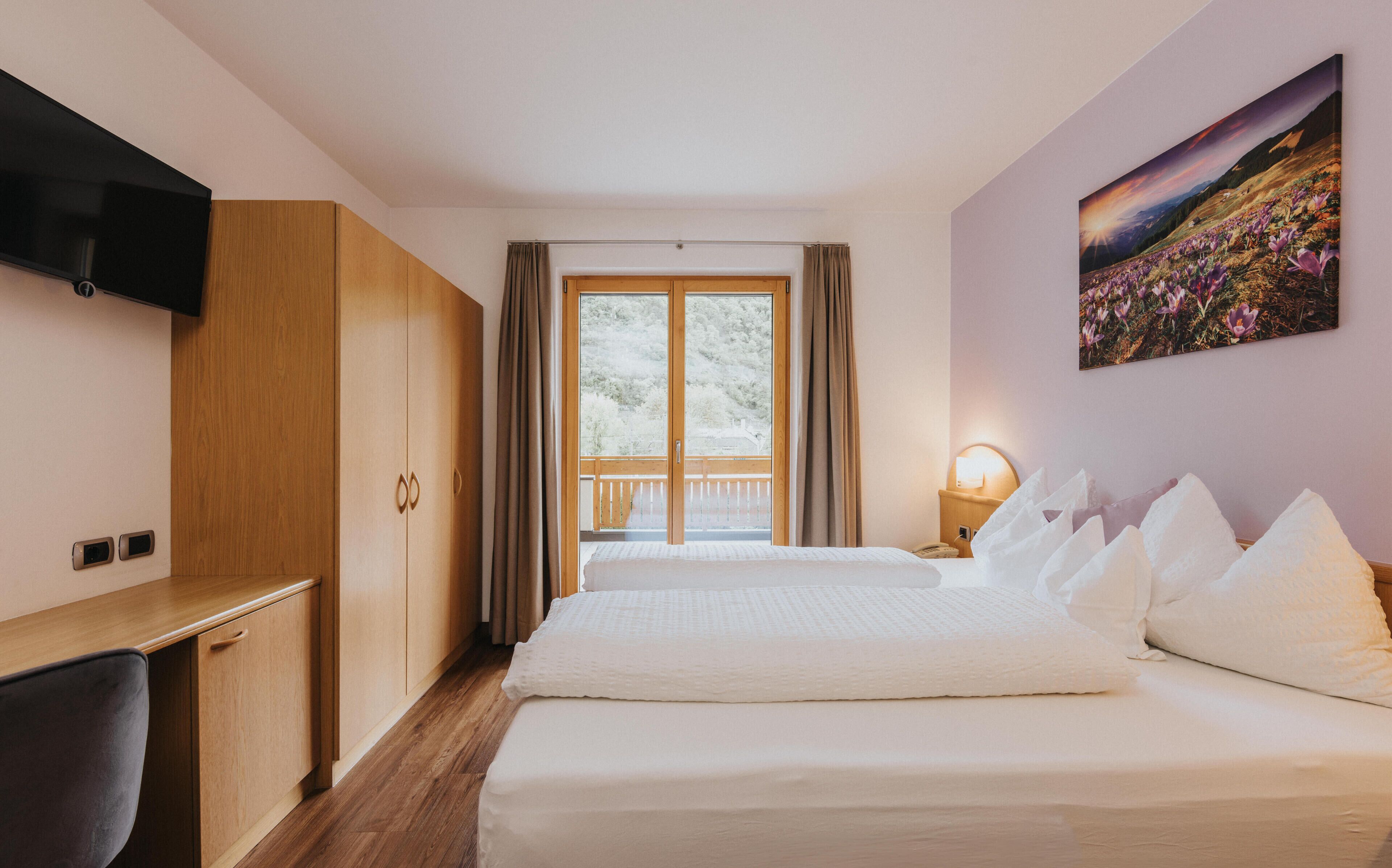 A hotel room with a bed, a desk, a TV, a window with curtains, and a painting on the wall.