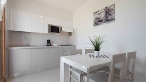 A modern kitchen with white cabinets, a microwave, and a coffee maker. There is a dining table with chairs and a potted plant.