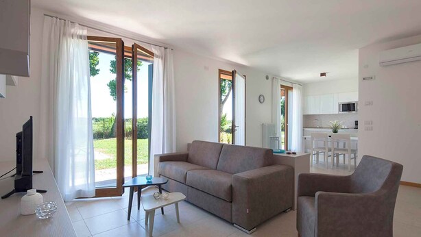 A modern living room with a sofa, a coffee table, and a TV. There is a dining area with a table and chairs. The room has large windows with curtains.