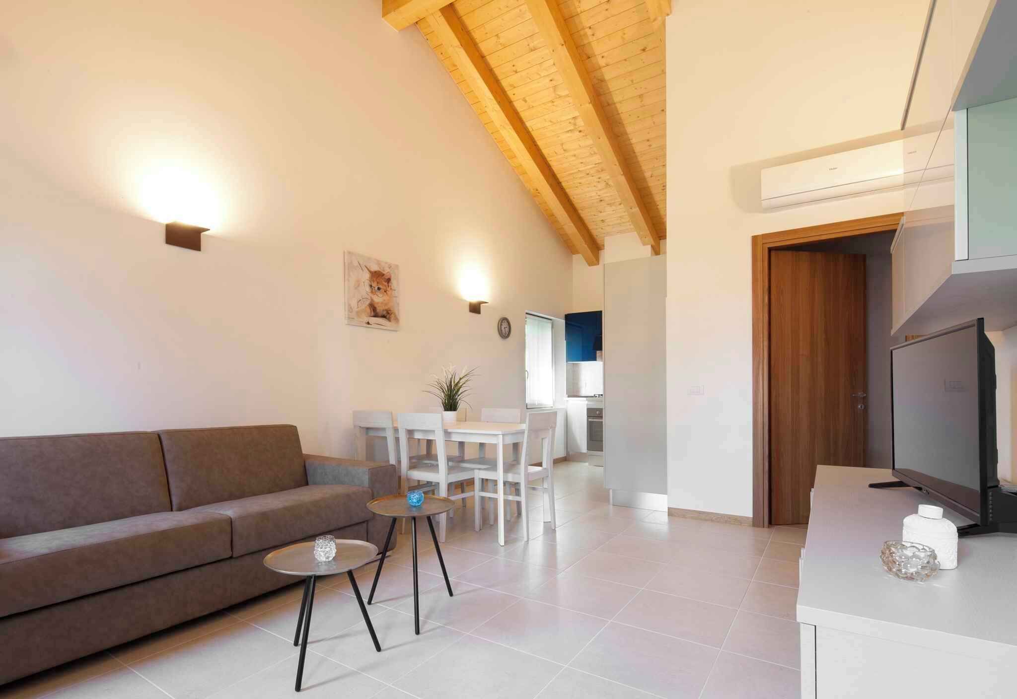 Una sala de estar moderna con sofá, mesa de centro y televisor. Hay un comedor con sillas y una mesa. La habitación tiene vigas de madera en el techo y una cocina al fondo.
