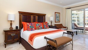 A hotel room with a large bed, a wooden nightstand, a desk, a chair, and a balcony with a view of buildings. - One Bedroom Suite (Cabo San Lucas)