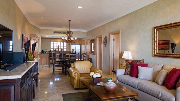 A spacious living room with a sofa, coffee table, and a dining area with chairs and a chandelier. - One Bedroom Suite (Cabo San Lucas)