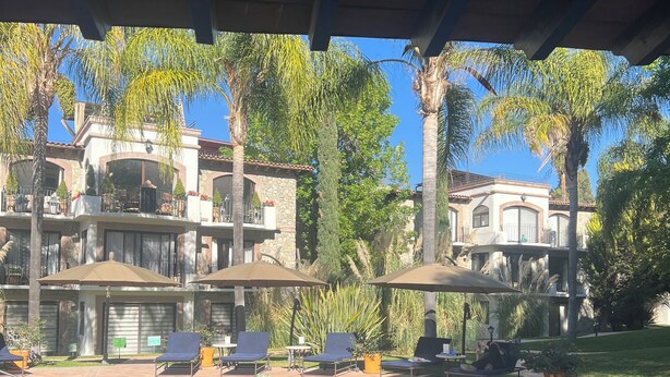 Un área de piscina con sillones de descanso y sombrillas, rodeada de palmeras y un edificio grande con varios balcones.