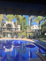 Un área de piscina con sillones de descanso y sombrillas, rodeada de palmeras y un edificio grande con varios balcones.
