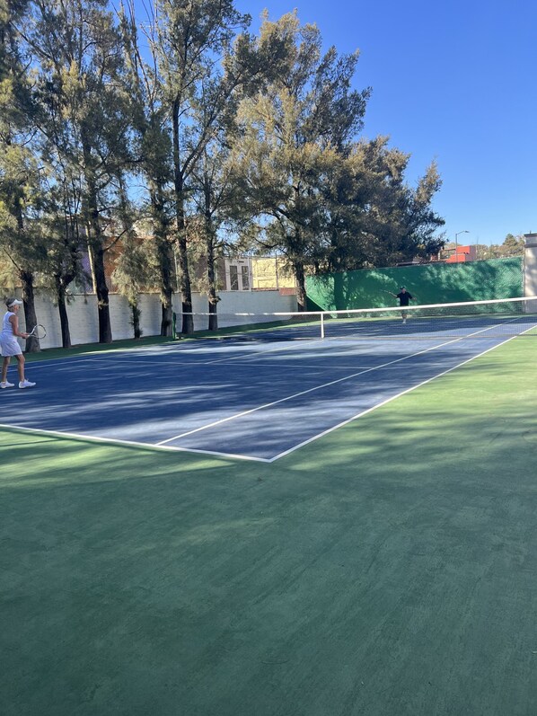 An outdoor tennis court with a net, surrounded by trees and a green fence.