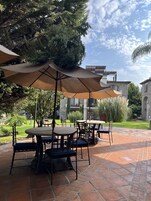 Un patio al aire libre con mesas y sillas bajo grandes sombrillas, rodeado de vegetación y edificios de piedra.