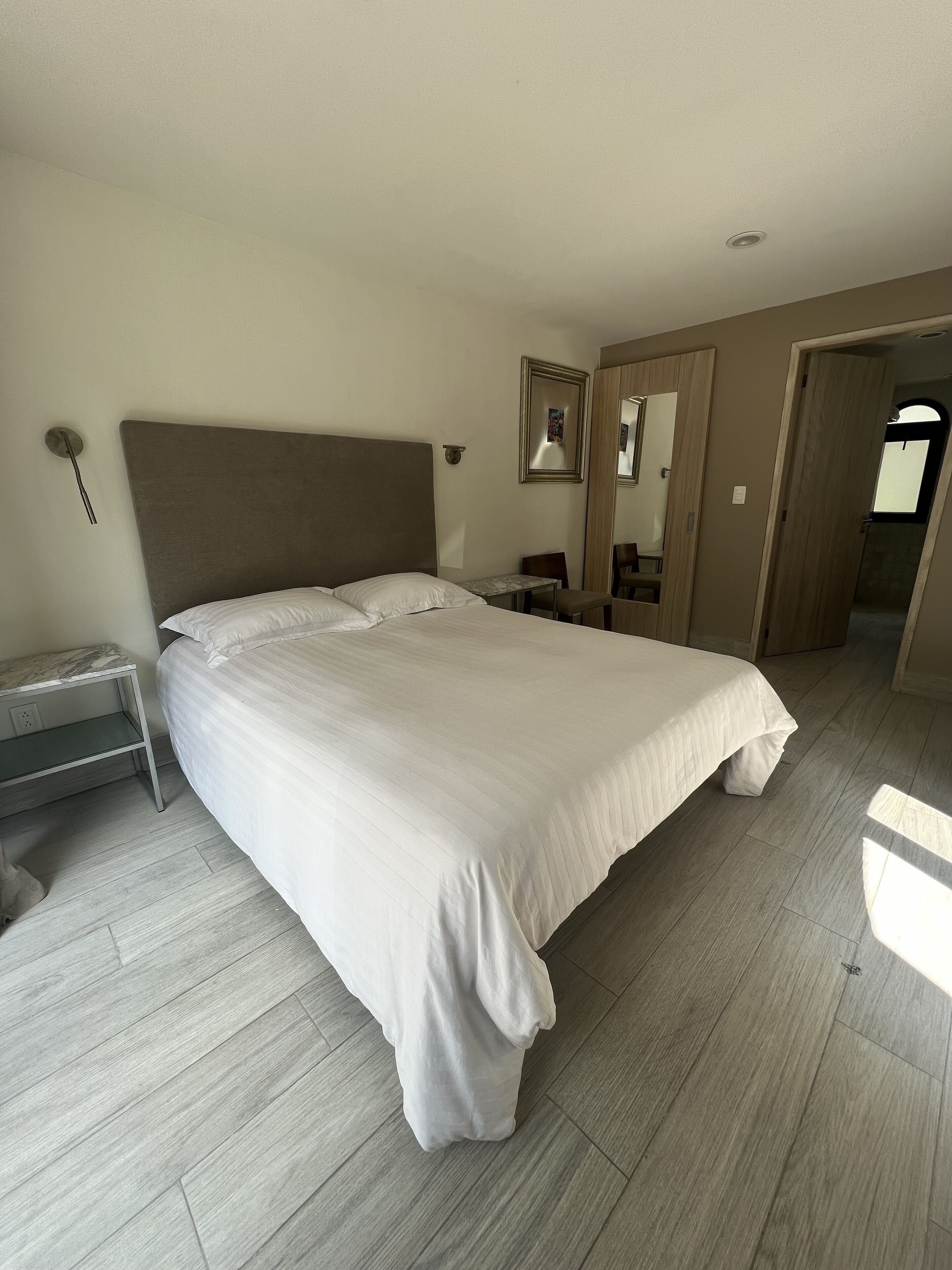 A modern hotel room with a large bed, a bedside table, a chair, and a mirror.