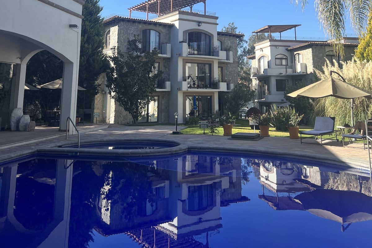 Vista de la piscina con un edificio de varios pisos con balcones, un cielo azul despejado y follaje circundante.