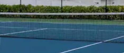 A tennis court with a net, surrounded by a fence and greenery.