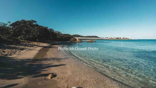 A beach with clear water, a sandy shore, and a distant view of buildings. - Ocean Escape - pools, beach, multiple courts, gym (Lucea)