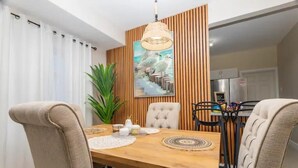 A dining area with a wooden table, beige upholstered chairs, a hanging light fixture, and a wall-mounted painting. - Ocean Escape - pools, beach, multiple courts, gym (Lucea)
