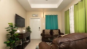 Un salon avec un canapé en cuir marron, une console en bois foncé sur laquelle se trouve un téléviseur, et un rideau vert.