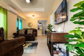 A living room with a brown sofa, a coffee table, and a TV on the wall. There is a plant on the right side and a bookshelf with books and decorative items.