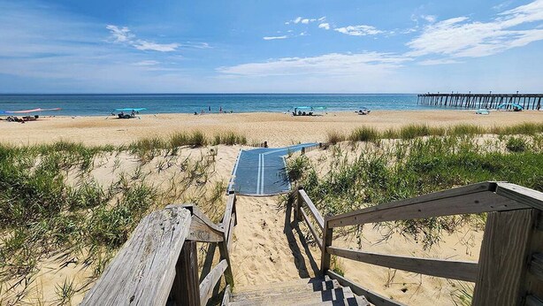 A beach with a wooden walkway leading to the water, a pier in the distance, and beachgoers. - Beautiful semi-oceanfront townhouse near Kitty Hawk Pier! (Kitty Hawk)