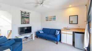 A living room with a TV, blue sofa, and a small kitchenette. - Beautiful semi-oceanfront townhouse near Kitty Hawk Pier! (Kitty Hawk)
