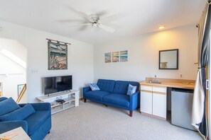 A living room with a TV, blue sofa, and a small kitchenette. - Beautiful semi-oceanfront townhouse near Kitty Hawk Pier! (Kitty Hawk)
