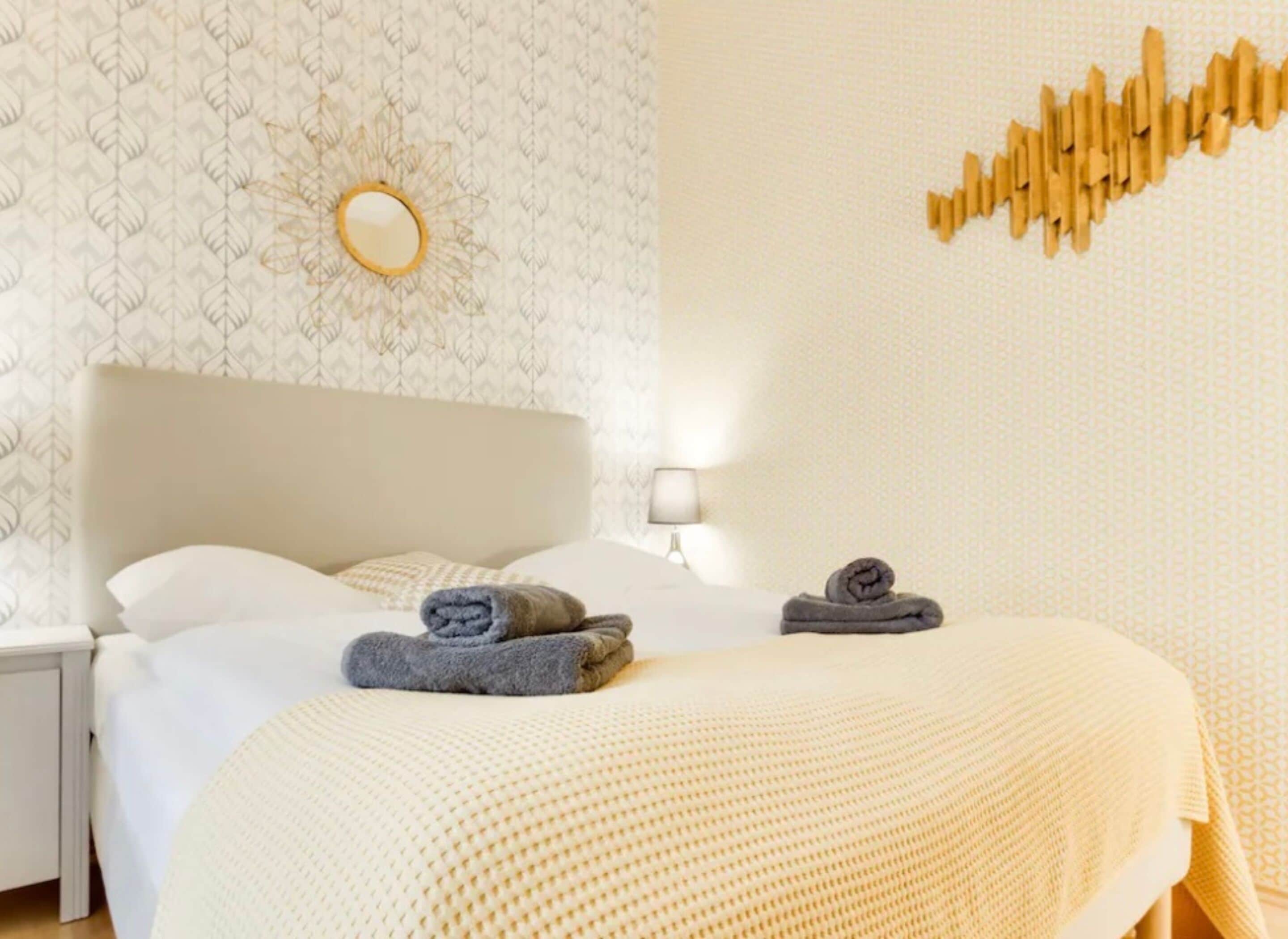 A neatly made bed with two rolled towels, a bedside lamp, and a wall-mounted mirror.