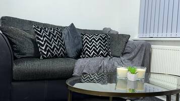 Uma sala de estar moderna com um sofá preto, almofadas estampadas, uma mesa de centro de vidro e um radiador.