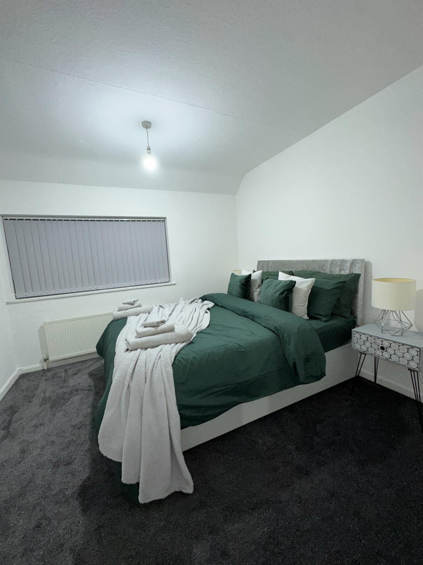 Quarto com cama, mesa de cabeceira, abajur, radiador e janela com persianas.