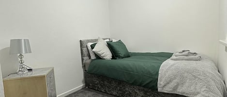 A small, single-bed room with a green bedspread, a bedside lamp, and a wooden nightstand.