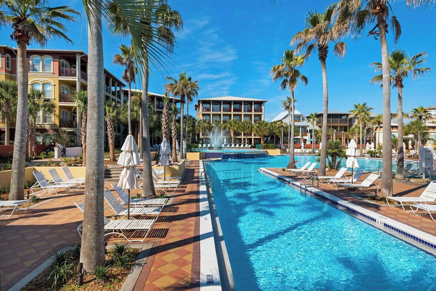 Una piscina con sillas de descanso y palmeras, rodeada de edificios de hotel.