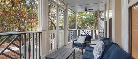 A covered porch with a wooden deck, a dark blue sofa with patterned cushions, and a ceiling fan.